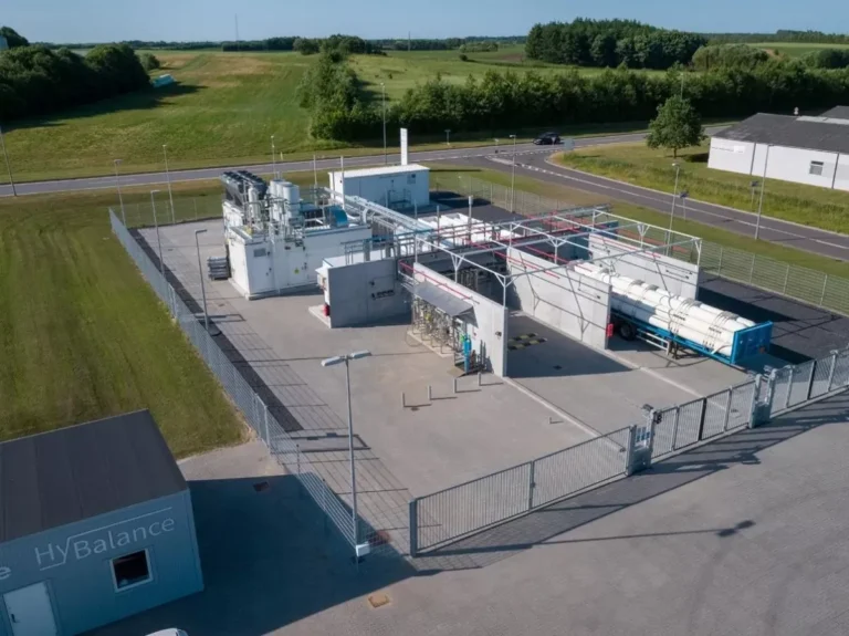 Aerial view of a fenced industrial hydrogen plant with white buildings, large storage tanks, and pipelines, situated next to a road and surrounded by green fields and trees.