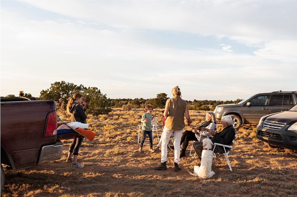 A group of people and a dog gather outdoors between two parked vehicles in a dry, grassy landscape. Some sit in folding chairs whilst others stand and talk, enjoying a relaxed moment under a partly cloudy sky.