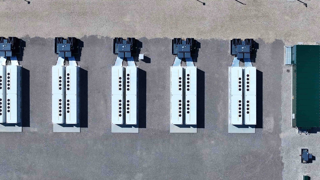 Aerial view of six large rectangular industrial units aligned in parallel on a gravel car park, with a small green-roofed building on the right side of the image.