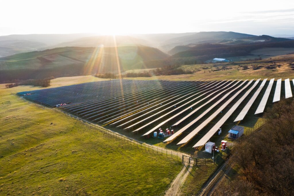 Aerial view of a large solar farm with rows of solar panels in a field, surrounded by hills and bathed in sunlight, with a few small buildings and equipment visible near the panels.