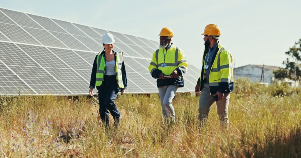 Three workers in safety gear and helmets walk through a grassy field next to large solar panels, discussing or inspecting the solar energy installation under a clear sky.