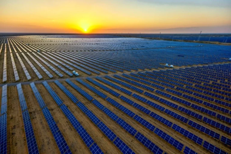 Aerial view of a vast solar farm with rows of solar panels stretching into the distance at sunset, capturing the sunlight on a flat landscape.